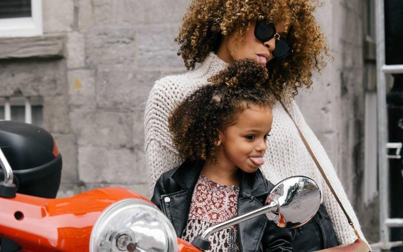 mother and daughter stick their tongues out while looking at themselves in a motorcycle mirror parked in front of a building